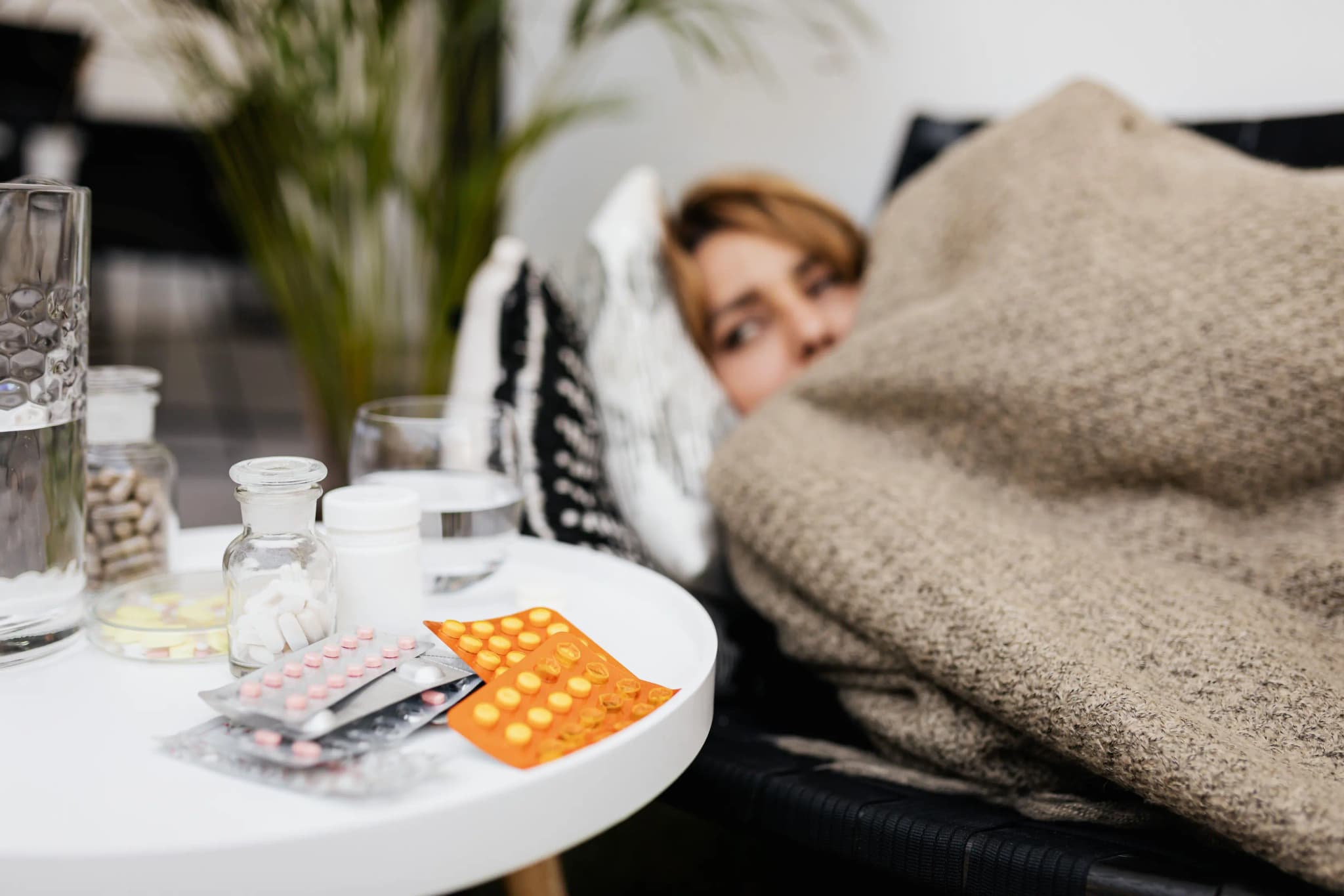 Eine Frau ruht unter einer Decke auf dem Sofa; Wasser und Medikamente stehen auf dem Tisch