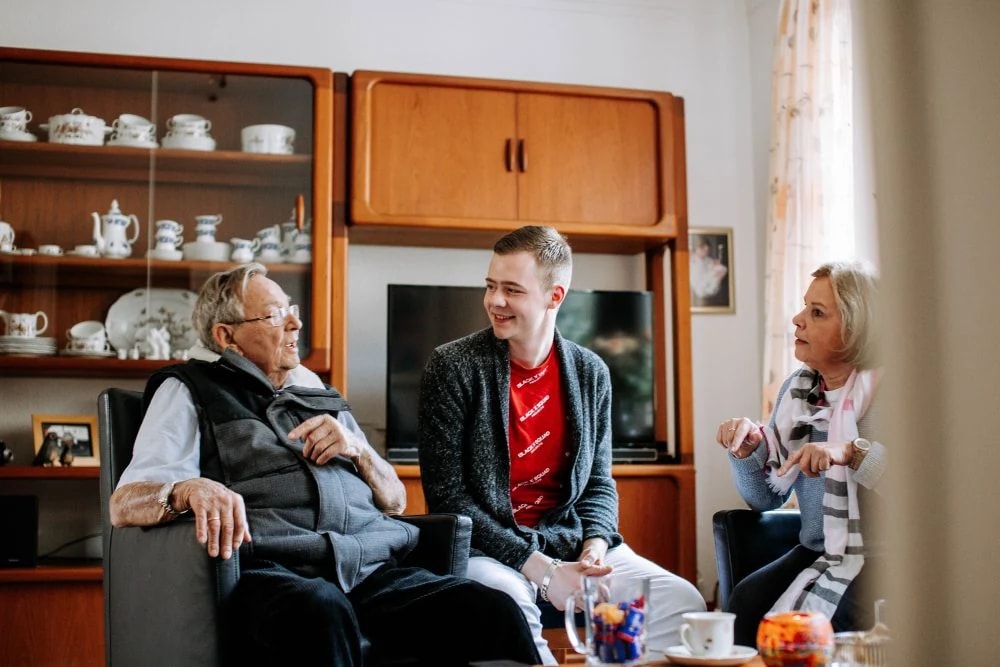 Eine Haushaltshilfe im Gespräch mit einem älteren Paar im Wohnzimmer.