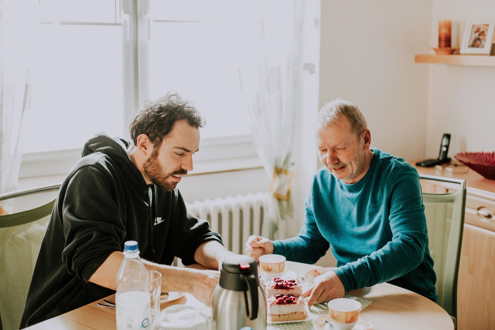 Ein junger Alltagsbegleiter und ein älterer Mann sitzen zusammen am Küchentisch bei Kaffee und Kuchen.