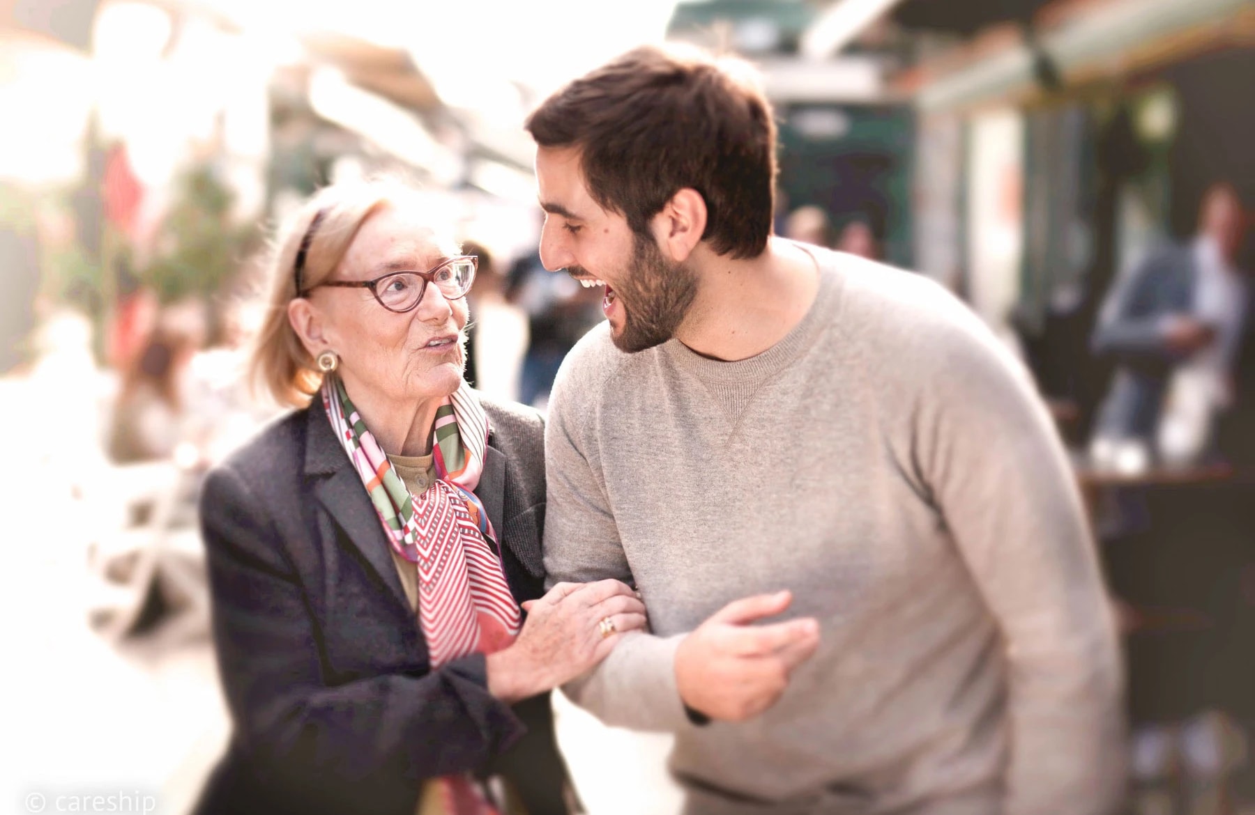 Eine Seniorin und ihr Alltagshelfer lachen gemeinsam bei einem Spaziergang über den Markt.