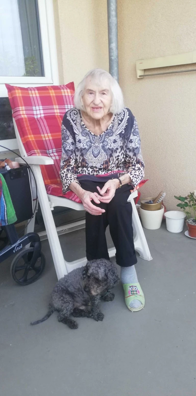 Ältere Frau sitzt auf ihrem Balkon, zu ihren Füßen liegt ein Hund